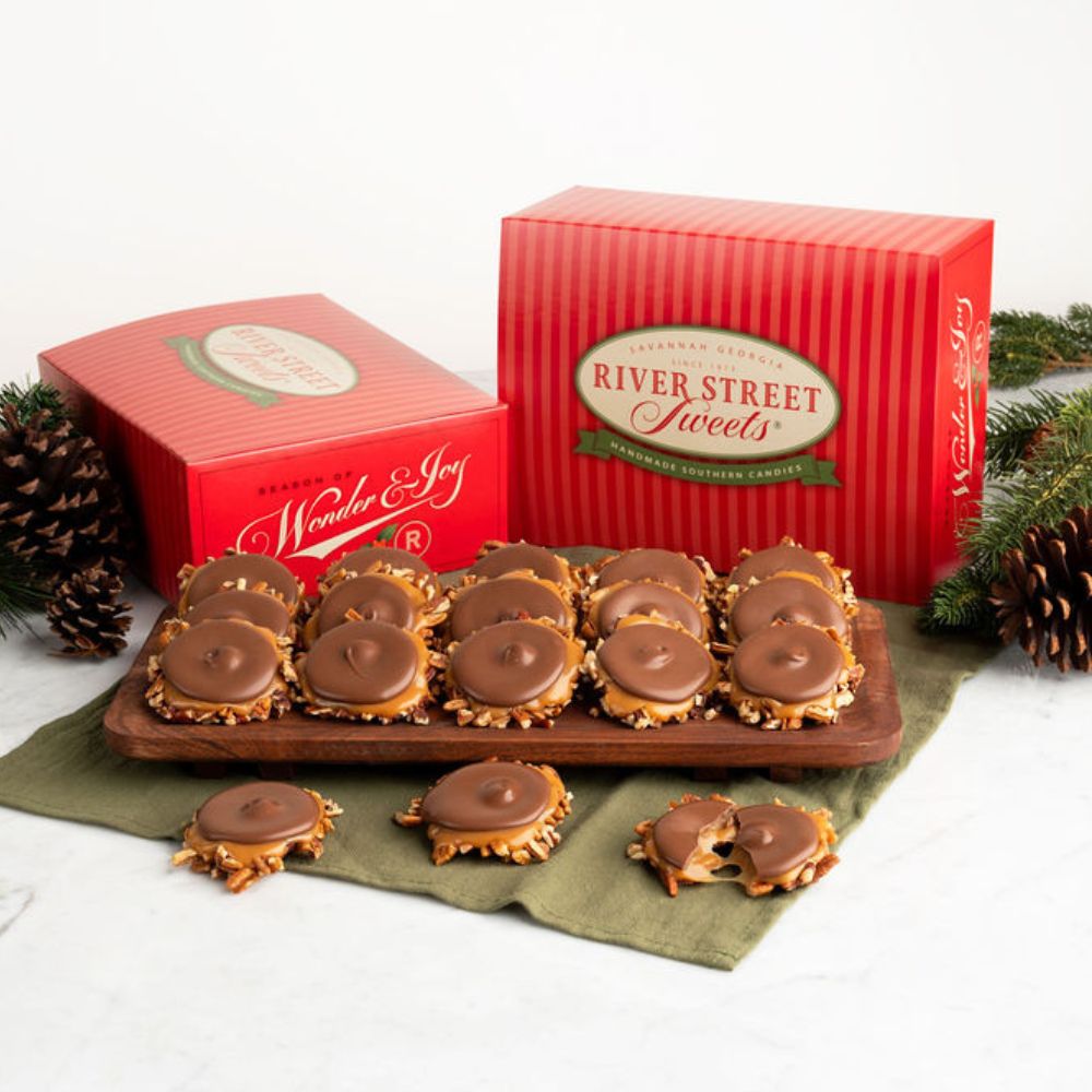 River Street Sweets chocolate bear claws displayed on a wooden tray with two red branded gift boxes, pinecones, and greenery on a light neutral background.