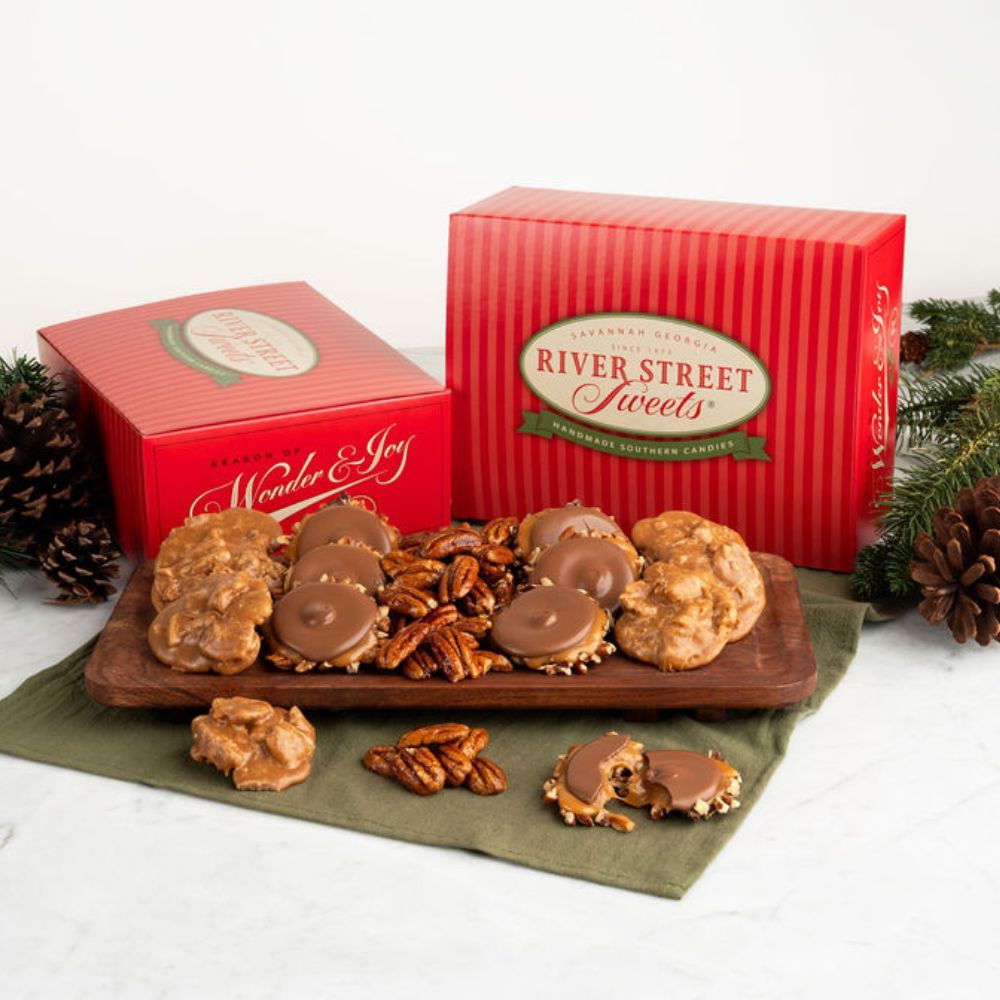A holiday assortment of River Street Sweets displayed on a wooden board with pecans, alongside two red gift boxes. The scene is styled with pinecones and evergreen branches on a white marble surface.