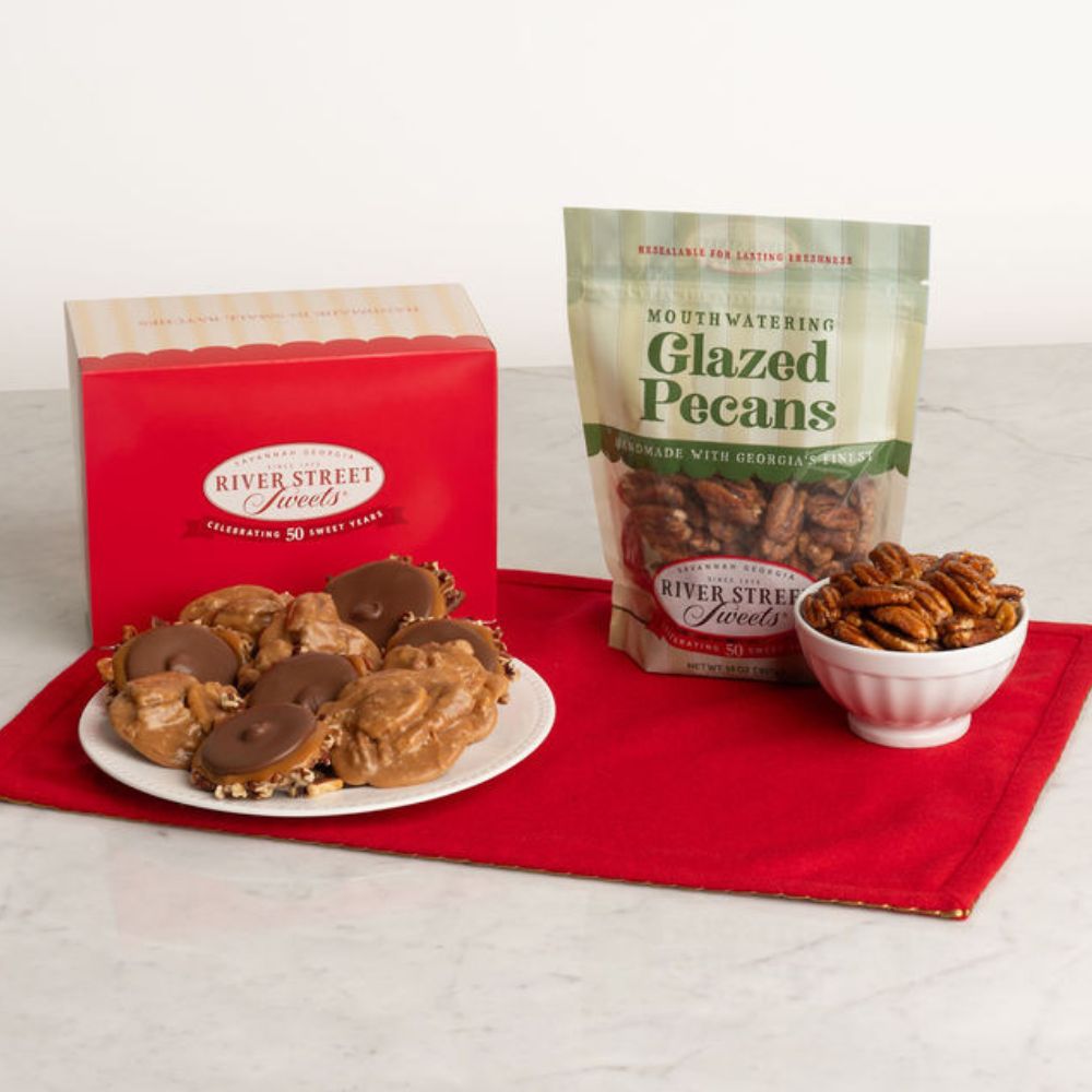 River Street Sweets gift set featuring a red box of pralines and bear claws, a bag of glazed pecans, and a small bowl of pecans arranged on a red cloth over a white marble surface.