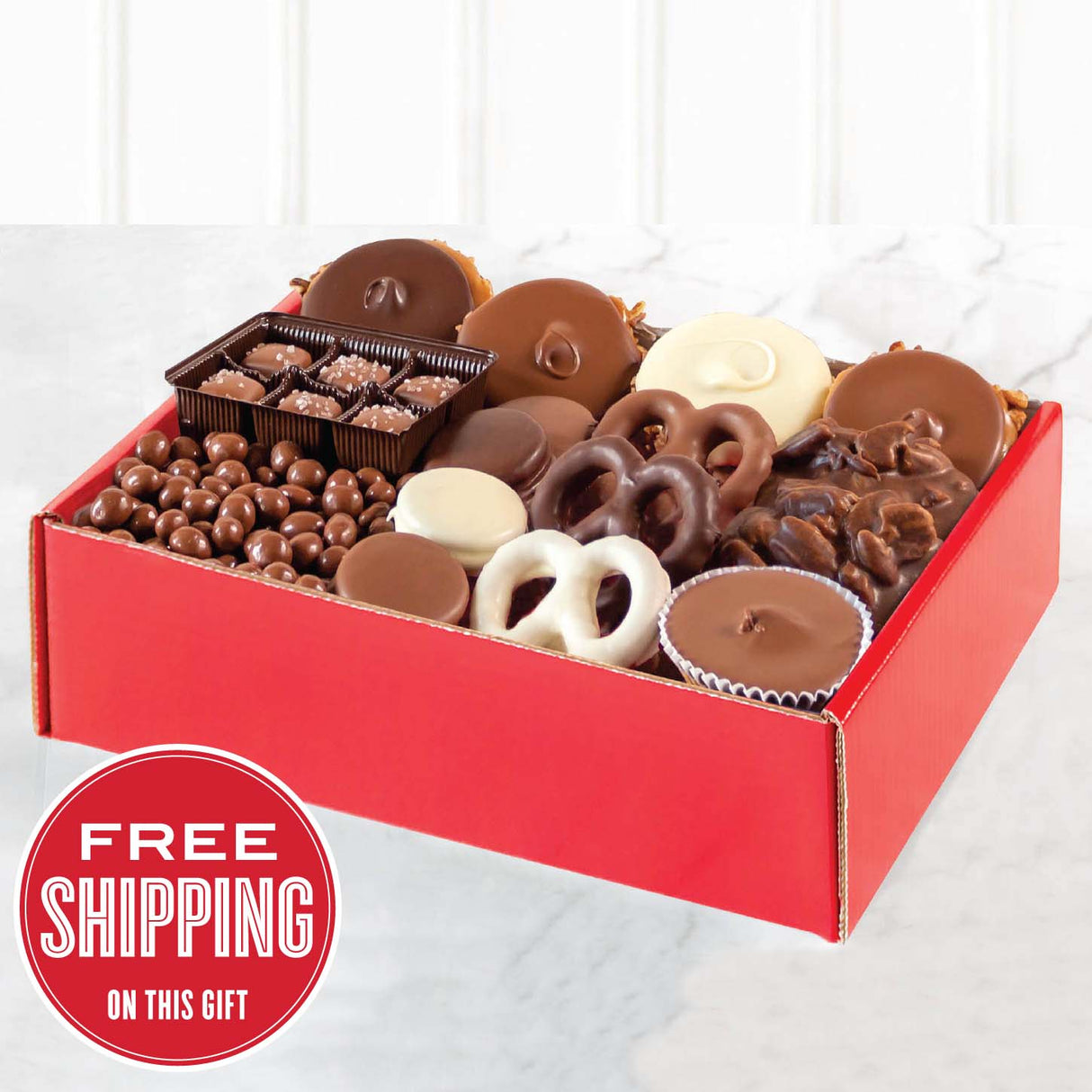 A red gift box filled with an assortment of chocolate candies including pralines, bear claws, a peanut butter cup, assorted pretzels, oreos, chocolate-covered peanuts, and sea salt caramels.