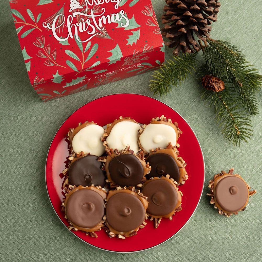 A styled holiday product shot showing a red plate of assorted bear claws, with a red Christmas gift box in the background and pine branches and pinecones on a green surface.