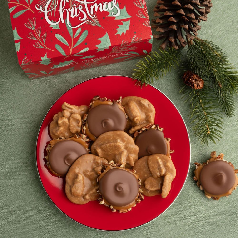 A top-down holiday flat lay showing a red plate filled with pralines and bear claws, with a red Christmas gift box, pinecones, and evergreen branches on a green textured surface.