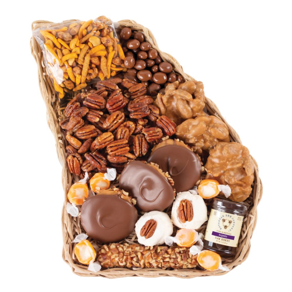 A wicker basket filled with assorted gourmet snacks and candies, including chocolate-dipped chocolates, pecans, peanuts, caramel-coated pieces, pretzel chips, and a small jar, photographed on a white background.