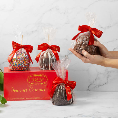 Gourmet caramel apples with red bows in front of a River Street Sweets box on a marble surface.