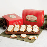 White chocolate bear claw assortment displayed on a wooden board with two red River Street Sweets gift boxes, pinecones, and evergreen branches on a light marble surface.