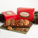 A holiday assortment of River Street Sweets displayed on a wooden board with pecans, alongside two red gift boxes. The scene is styled with pinecones and evergreen branches on a white marble surface.