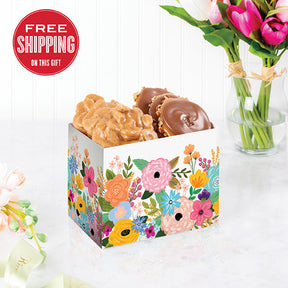 A box of floral patterns containing chocolate bear claws and pralines, placed on a white surface with a vase of pink tulips in the background.