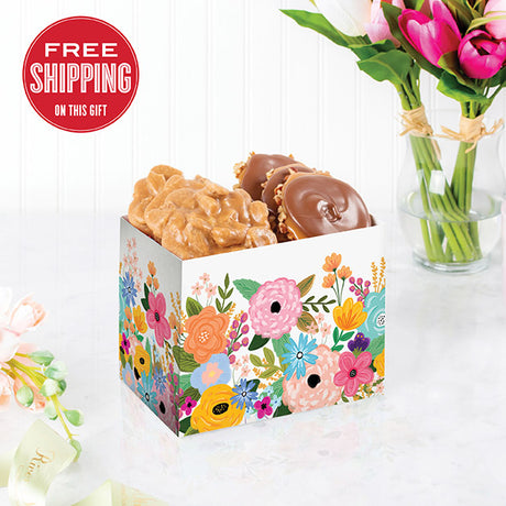 A box of floral patterns containing chocolate bear claws and pralines, placed on a white surface with a vase of pink tulips in the background.