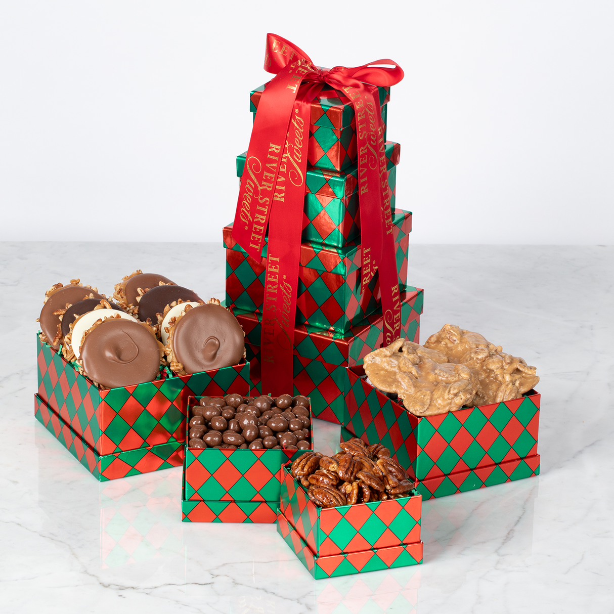 A festive holiday gift tower with red and green boxes, containing various chocolate treats and nuts, adorned with a red ribbon.