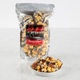 A clear plastic package of Savannah Crunch snack, showing crunchy caramel popcorn with pecans and chocolate drizzle. A small white bowl with the same snack is also presented in front of the package.