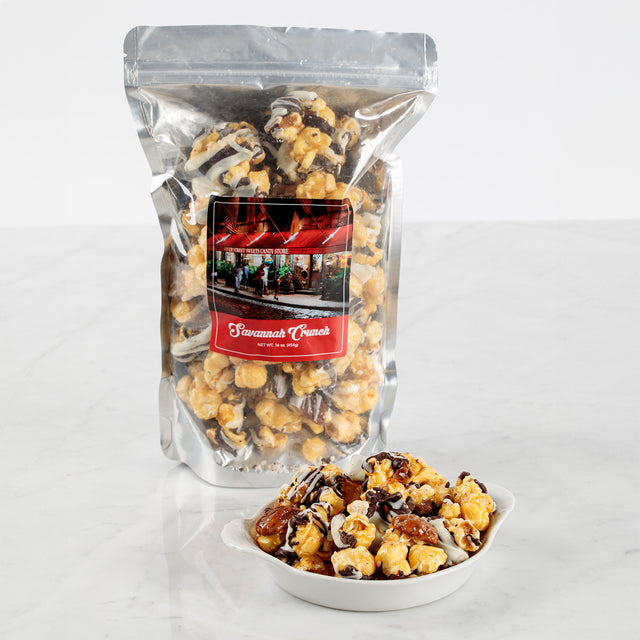 A clear plastic package of Savannah Crunch snack, showing crunchy caramel popcorn with pecans and chocolate drizzle. A small white bowl with the same snack is also presented in front of the package.
