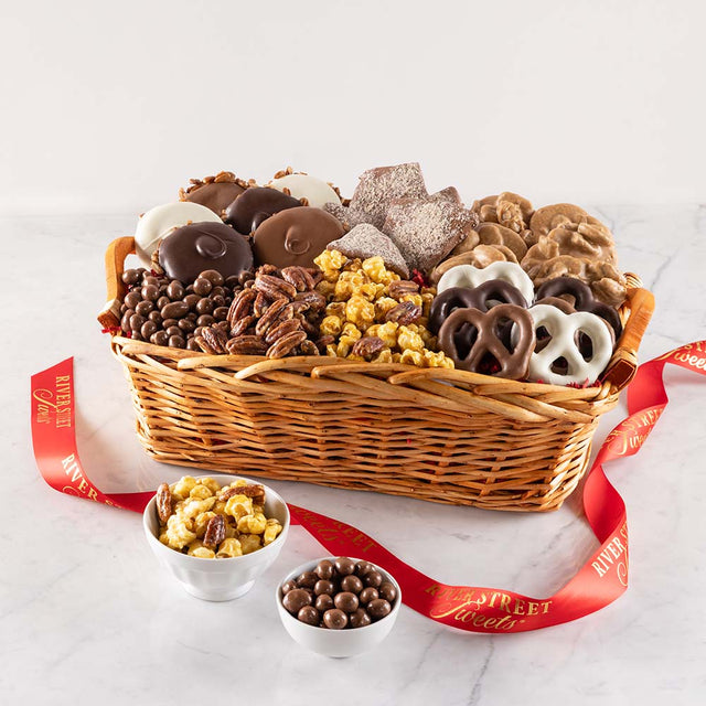 A wicker gift basket filled with an assortment of sweet treats including pralines, chocolate covered pretzels, glazed pecans, and other confections, with wooden handles on the sides.