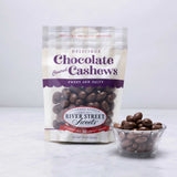 A packaged bag of River Street Sweets Chocolate Covered Cashews with a transparent section showing the cashews inside. There is also a small bowl of chocolate covered cashews beside the bag.