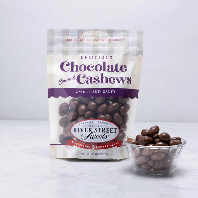 A packaged bag of River Street Sweets Chocolate Covered Cashews with a transparent section showing the cashews inside. There is also a small bowl of chocolate covered cashews beside the bag.