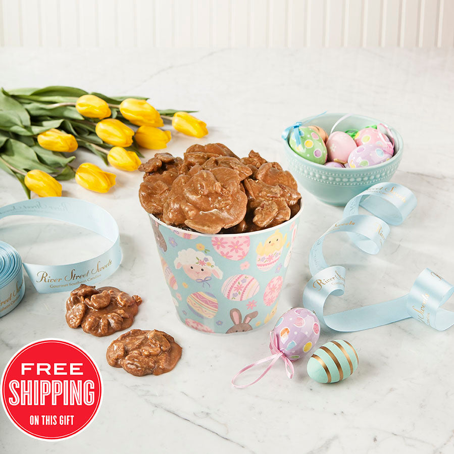 A colorful Easter-themed pail filled with individually wrapped pralines. The pail has illustrations of eggs and bunnies, and is accompanied by yellow tulips, multicolored Easter eggs, and decorative ribbons.