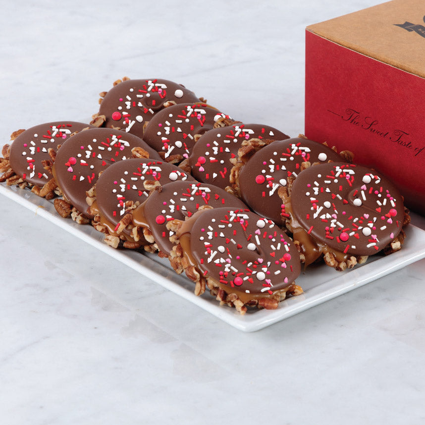A box of Valentine's Day Festive Sprinkle Bear Claws displayed on a white plate, consisting of chocolate covered bear claws with festive red and white sprinkles on top.