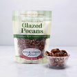 A resealable pouch of glazed pecans with some pecans visible in a small clear bowl beside it.