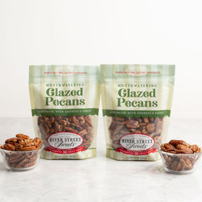 Two bags of River Street Mouthwatering Glazed Pecans displayed on a light marble surface with two small glass bowls of pecans in front, against a clean white background.