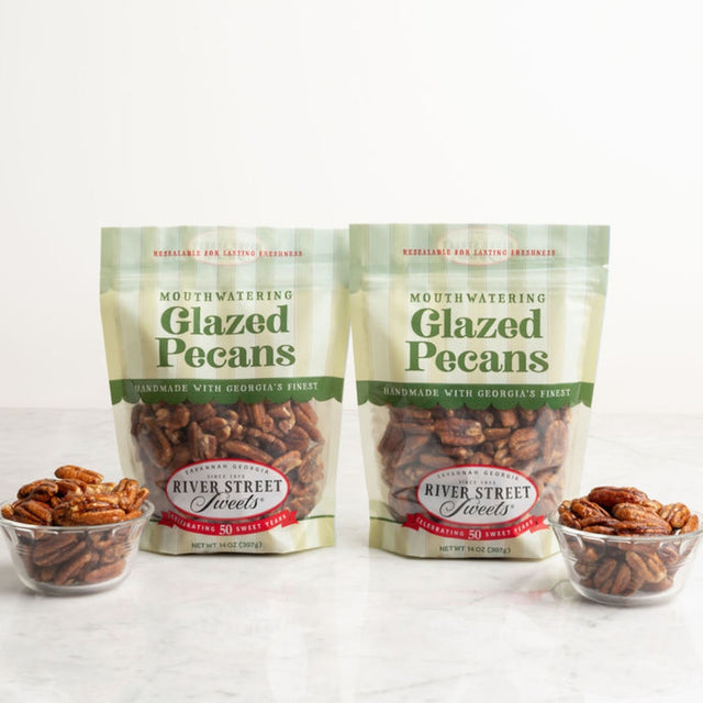 Two bags of River Street Mouthwatering Glazed Pecans displayed on a light marble surface with two small glass bowls of pecans in front, against a clean white background.