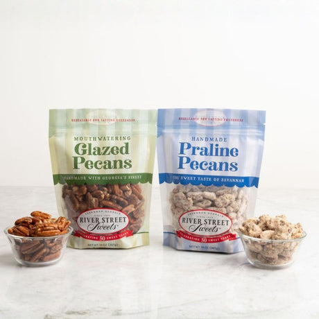 Two stand-up pouches of River Street Sweets handcrafted pecans on a light marble surface with two small glass bowls of pecans in front. The left pouch is labeled Glazed Pecans in pale green, and the right pouch is labeled Praline Pecans in light blue.