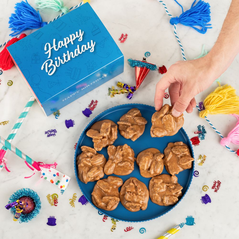 Hand picking a praline from a blue plate with a 'Happy Birthday' card and party decorations.