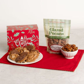 A holiday gift set featuring a red Christmas box of pralines and bear claws, a bag of Mouthwatering Glazed Pecans, and a small bowl of pecans arranged on a red cloth over a white marble surface.