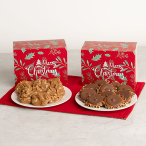 Two red Christmas-themed gift boxes with green holly patterns and white 'Merry Christmas' script, each placed behind a plate of holiday treats on a red cloth. The left plate holds pralines, and the right plate holds chocolate bear claws.