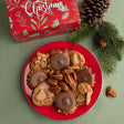 A red holiday-themed box with Christmas lettering sits behind a red plate filled with pralines, pecans, and bear claws, styled with pine branches and pinecones on a green surface.