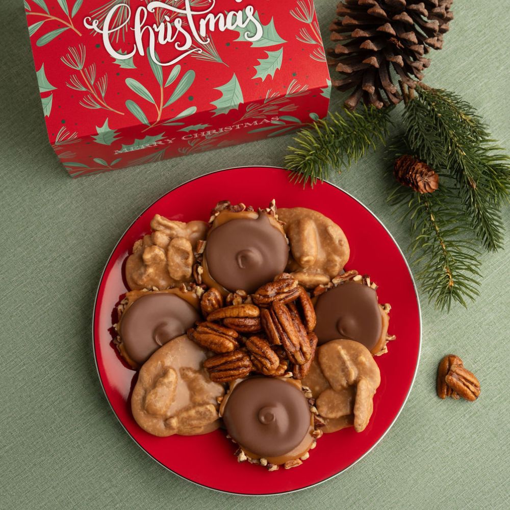 A red holiday-themed box with Christmas lettering sits behind a red plate filled with pralines, pecans, and bear claws, styled with pine branches and pinecones on a green surface.