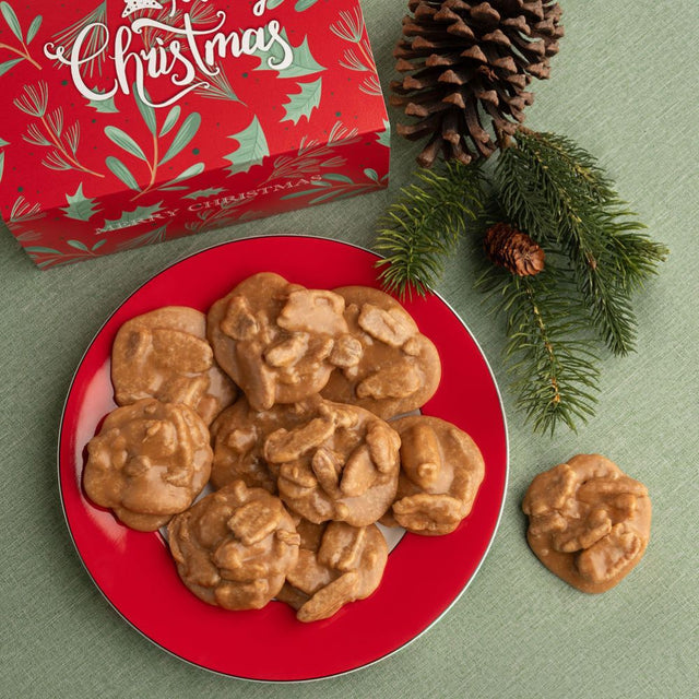 Pralines on a red plate next to a red Christmas box, styled with pinecones and evergreen branches on a green background.