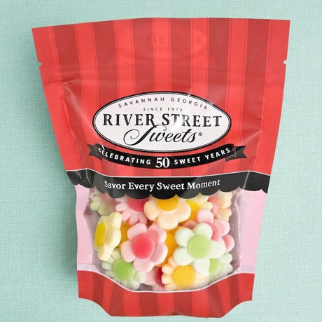 A bag of River Street Sweets flower-shaped gummy candies on a textured light blue background. The packaging is red and white with a clear window showing the colorful flower gummies inside.