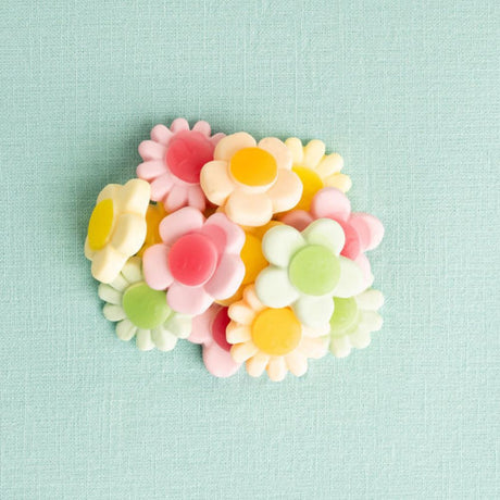 A cluster of small daisy-shaped candies in pastel pink, yellow, green, and white, arranged on a light mint green textured fabric background.