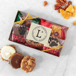 A clear gift box containing an assortment of chocolate bear claws and glazed nuts, tied with a red ribbon. A few pieces are placed outside of the box, and the brand's logo is visible on a tag within the box.