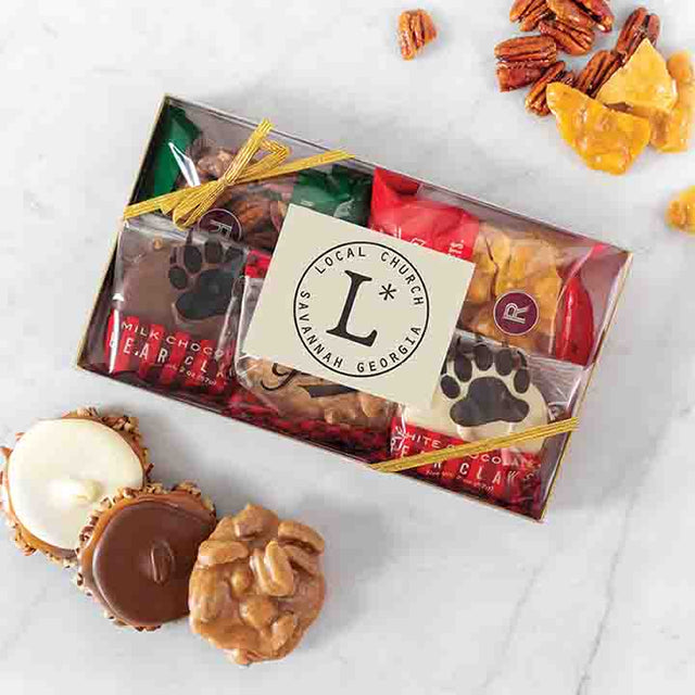 A clear gift box containing an assortment of chocolate bear claws and glazed nuts, tied with a red ribbon. A few pieces are placed outside of the box, and the brand's logo is visible on a tag within the box.