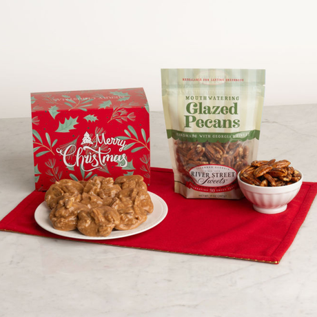 A holiday gift set featuring a red 'Merry Christmas' box, a bag of Mouth Watering Glazed Pecans by River Street Sweets, and a small bowl of pecans on a red cloth over a white marble surface.