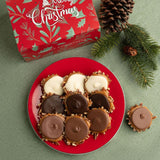 A styled holiday product shot showing a red plate of assorted bear claws, with a red Christmas gift box in the background and pine branches and pinecones on a green surface.