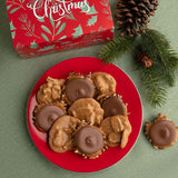 A top-down holiday flat lay showing a red plate filled with pralines and bear claws, with a red Christmas gift box, pinecones, and evergreen branches on a green textured surface.