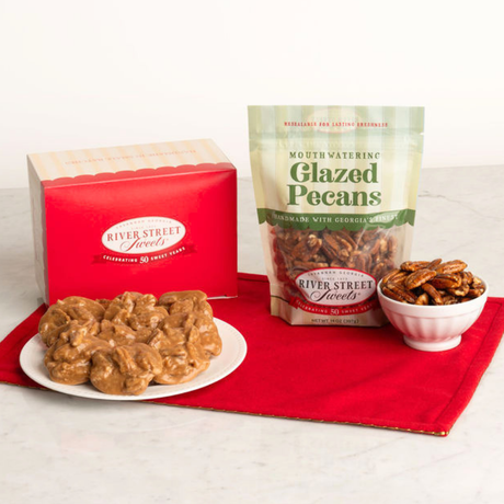 River Street Sweets glazed pecans displayed with a red gift box, a stand-up pouch of glazed pecans, and a small bowl of pecans on a red cloth over a light marble surface.