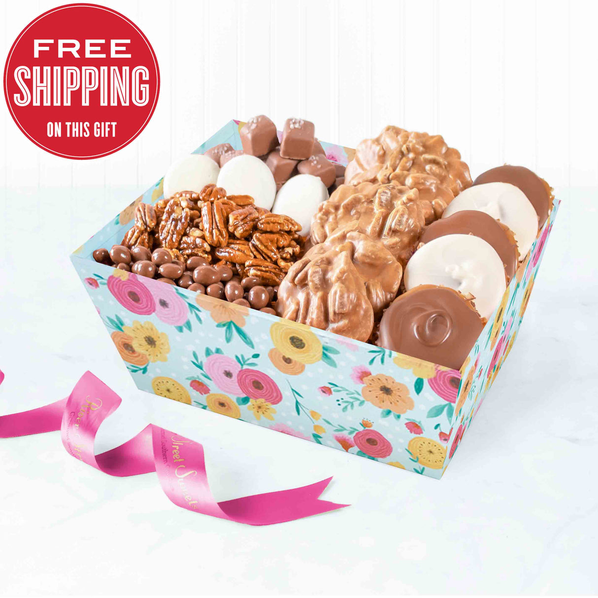 A gift tray filled with an assortment of chocolates including pralines, bear claws, white chocolate-dipped Oreos, glazed pecans, sea salt caramels, and chocolate covered almonds. The tray has a floral pattern and there is a pink ribbon next to it.