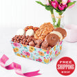 An assortment of chocolates and nuts presented in a floral gift box, with a 'Free Shipping on this gift' badge, and a pink ribbon. A vase with pink flowers and a pink cup are in the background.