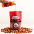 A red stand-up pouch of River Street Sweets glazed pecans sits on a white surface surrounded by scattered pecans, with a hand pouring honey from an orange bottle into the pouch.