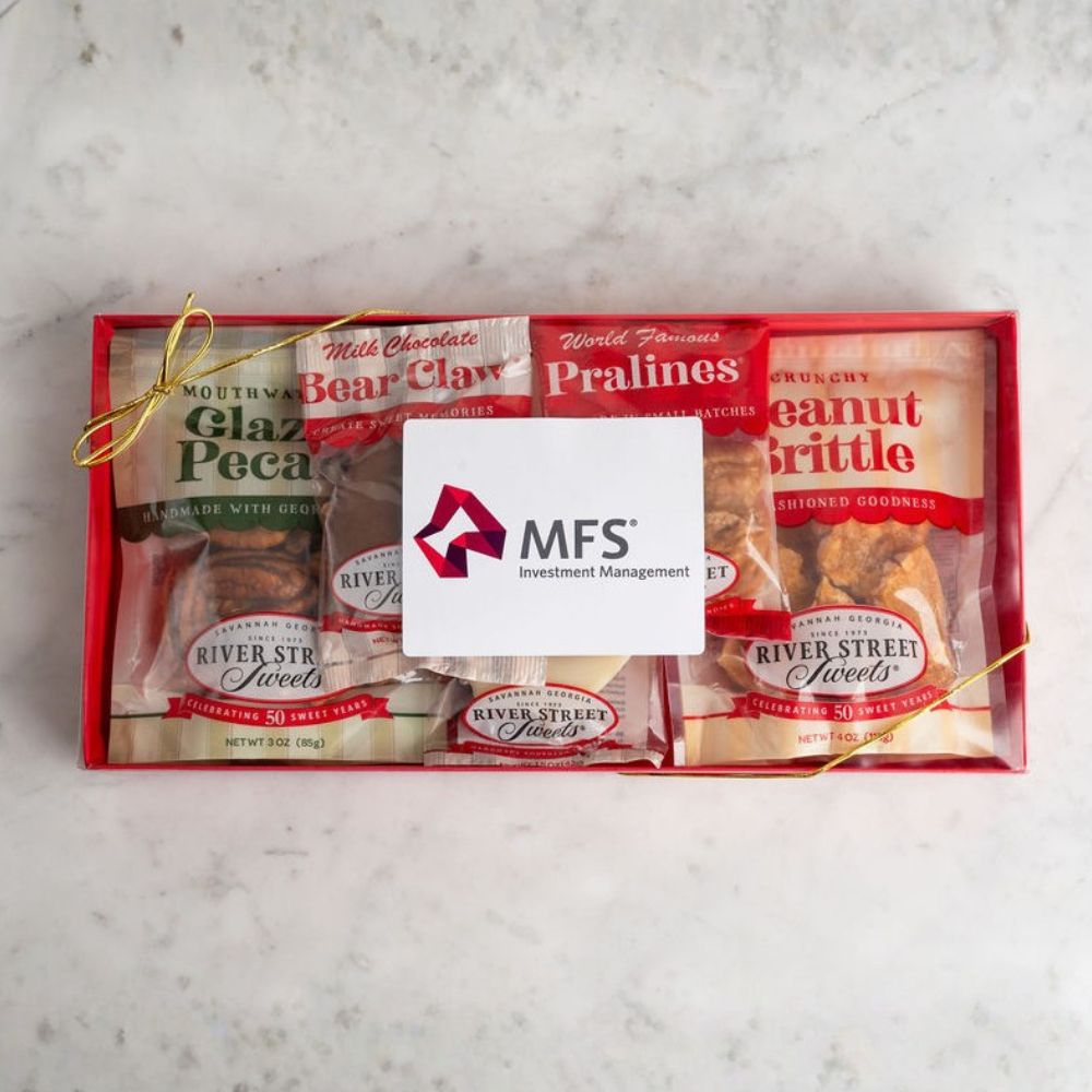 A gift box of gourmet snacks and sweets arranged in a red tray, tied with a gold string and covered with a white card. Visible packaged items include Chocolate Bear Claw, World Famous Pralines, Peanut Butter Brittle, and River Street Sweets.