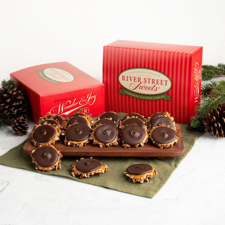 A set of holiday dark chocolate bear claws displayed on a wooden board with two red gift boxes from River Street Sweets, styled with pinecones and evergreen branches on a white surface.