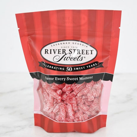 A bag of River Street Sweets candy on a light marble surface. The packaging is red and white with black accents, featuring the brand logo and a clear window showing the pink-red candy pieces inside.