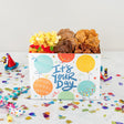 Gift box with snacks and a card reading 'It's Your Day' on a confetti-covered surface.