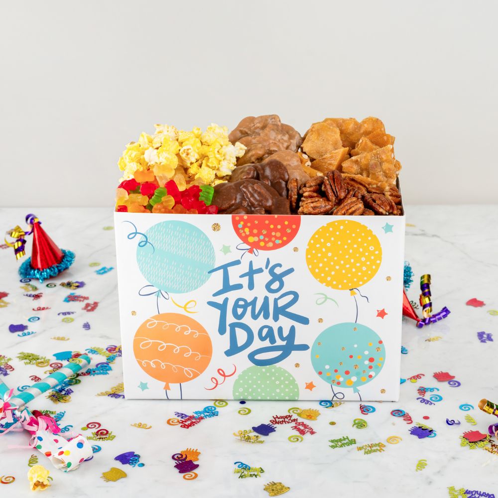 Gift box with snacks and a card reading 'It's Your Day' on a confetti-covered surface.