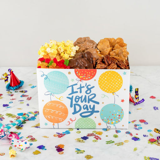Gift box with snacks and a card reading 'It's Your Day' on a confetti-covered surface.