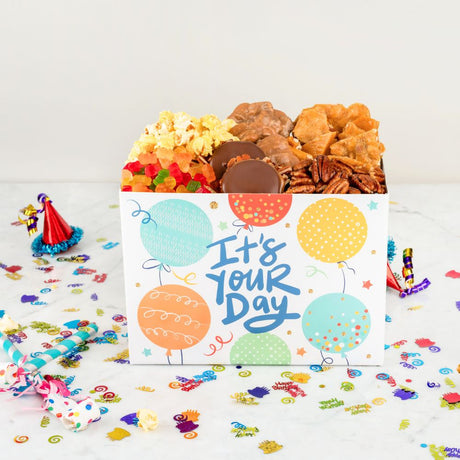 Gift box with snacks and a card saying 'It's Your Day' on a white background with confetti.