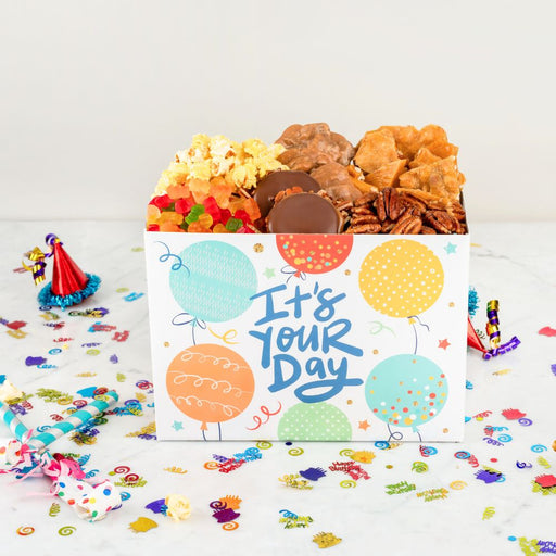 Gift box with snacks and a card saying 'It's Your Day' on a white background with confetti.