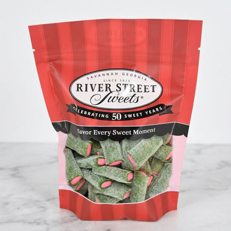 A bag of River Street Sweets candy on a light marble surface against a white background. The packaging is red and white striped with a clear window showing green sugar-dusted candy pieces with pink accents.